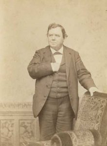 Sepia-toned image of Donnelly in a suite standing near a chair