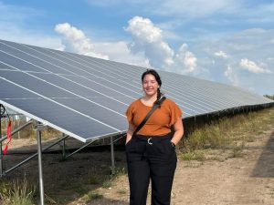 Sarah LaVallie standing outside in front of a solar panel on a bright sunny day, smiling and looking at the camera