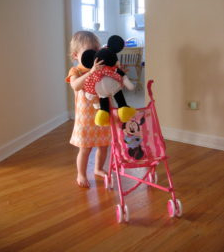 A young girl holds a stuffed animal while pushing a stroller.