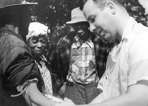 A white doctor administers an injection to a black participant in the Tuskegee Syphilis Study while two others look on.