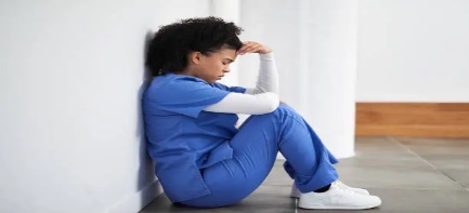 A person in blue scrubs sitting on the floor with her head in her hands looking down appearing stressed and burned out