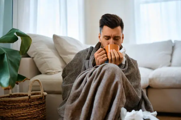 A person sitting on the floor wrapped in a blanket drinking from a mug with tissues around him appearing sick