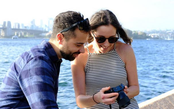 A young adult couple look at a camera screen together as if looking at a picture they have just taken of the scenery around them.